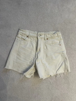 Krämvita jeansshorts från Zara - Snygga krämvita jeansshorts från Zara med råa, fransiga benslut och klassisk femficksdesign. Shortsen har hög midja och bälteshällor, perfekta för en avslappnad och trendig sommarlook.