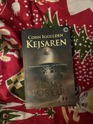 Kejsaren: Roms portar av Conn Iggulden - En episk roman av Conn Iggulden om antikens Rom, där gladiatorer, politiska intriger och krig formar berättelsen om Julius Caesars uppväxt. Omslaget är mörkt med guldtext och en hjälm i centrum. Perfekt för dig som gillar historiska äventyr.