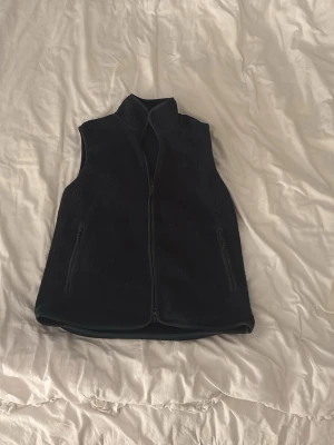 Mörkblå fleeceväst från Peak Performance - Mysig mörkblå fleeceväst från Peak Performance med hel dragkedja framtill och två praktiska fickor med dragkedja. Västen har hög krage och diskret broderad logga på bröstet. Perfekt för lager-på-lager och outdoor-vibes.