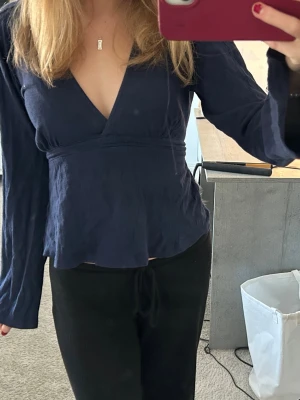 Mörkblå omlottopp med v-ringning - Snygg mörkblå omlottopp med v-ringning och långa ärmar. Toppen har en markerad midja och lätt utsvängd nederdel, vilket ger en smickrande siluett. Perfekt för dig som gillar stilrena och trendiga plagg.