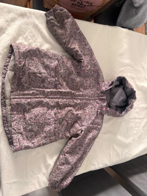 Blommig vindjacka med huva - Snygg vindjacka med blommigt mönster i rosa och grå toner. Jackan har huva, dragkedja framtill och två fickor. Perfekt för blåsiga dagar och enkel att matcha till olika outfits.