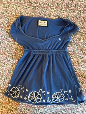 Blå babydoll topp med blommor från Abercrombie - Söt blå babydoll topp från Abercrombie med v-ringning och broderade vita blommor längs nederkanten. Toppen har korta ärmar och resår i midjan 💗💗💗
