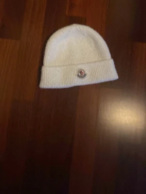 Vit ribbstickad mössa från Moncler - Säljer en stilren vit ribbstickad mössa från Moncler med uppvikt kant och klassisk Moncler-logga framtill. Perfekt accessoar för kalla dagar och enkel att matcha med olika outfits.