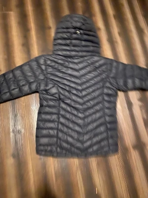 Svart dunjacka från Peak Performance - Snygg svart dunjacka från Peak Performance med huva och quiltad design. Jackan har dragkedja framtill och två sidofickor med dragkedja. Perfekt för kalla dagar och enkel att matcha med olika outfits.