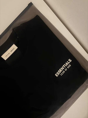 Svart Essentials Fear of God t-shirt - Snygg svart t-shirt från Essentials Fear of God med vit logga tryckt på bröstet. Klassisk rund hals och korta ärmar. Perfekt för dig som gillar streetwear och vill ha en stilren look. Tillverkad i mjuk bomull för maximal komfort.