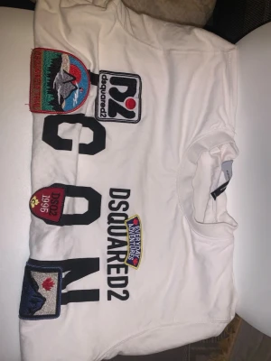 Vit DSQUARED2 t-shirt med patches - Säljer en vit t-shirt från DSQUARED2 med flera färgglada patches och tryck på framsidan. T-shirten har rund hals och korta ärmar. Stora bokstäver och coola märkesdetaljer gör den unik och trendig.