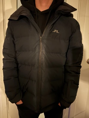 Svart pufferjacka från J.Lindeberg - Svart pufferjacka från J.Lindeberg med diskret logga på bröstet. Jackan har huva, dragkedja framtill och en klassisk quiltad design. Perfekt för kalla dagar med sin varma och vadderade känsla. Snygg och enkel att matcha till olika outfits.  OBS Lite solblekt framtill