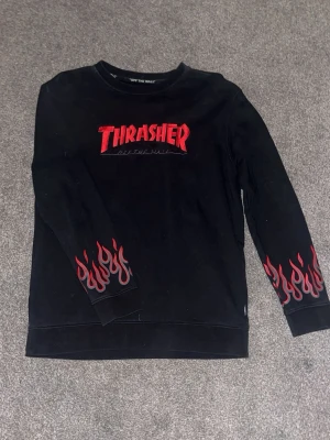 Thrasher x Vans  - Svart långärmad tröja från Thrasher med röd logga och texten 'Off The Wall' på bröstet. Ärmarna har röda flames tryckta vid mudden och baksidan har en liten Vans-logga upptill. Perfekt för dig som gillar streetwear och skate-stil.