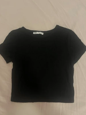 Svart croppad t-shirt från Zara - En enkel och stilren svart croppad t-shirt från Zara. T-shirten har rund halsringning och korta ärmar. Perfekt att matcha med höga jeans eller kjol för en clean look.