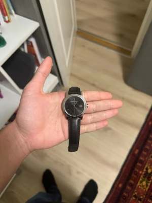 Burberry svart klocka med läderband - Stilren klocka från Burberry med svart urtavla och diskret rutmönster. Klockan har ett klassiskt svart läderband och silverfärgat boett. Datumvisning vid klockan tre och minimalistiska markeringar. Perfekt accessoar för en snygg och tidlös look. Need the money för studenten 