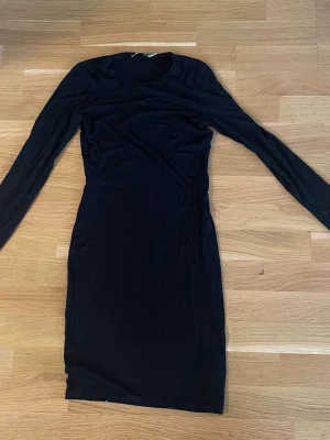 Svart ribbad långärmad klänning - En svart, ribbad klänning med lång ärm och rund hals. Klänningen är kroppsnära och har en kort längd, vilket ger en stilren och enkel look. Perfekt för dig som gillar minimalistisk stil.