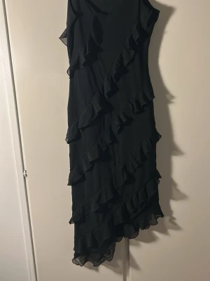 Svart volangklänning ärmlös - Snygg svart klänning med volangdetaljer i diagonala lager över hela klänningen. Ärmlös modell med asymmetrisk nederkant som ger en cool look. Perfekt för dig som vill sticka ut lite extra. Materialet känns lätt och följsamt.