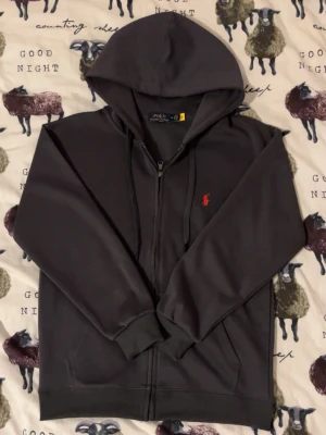  Polo Ralph Lauren - Snygg mörkgrå hoodie från Polo Ralph Lauren med dragkedja, huva och dragsnören. Klassisk röd broderad logga på bröstet och två fickor framtill. Tillverkad i mjuk bomullsmix som känns skön mot huden. Perfekt för en avslappnad och stilren look.