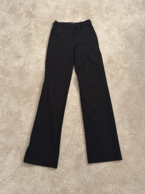 Svarta kostymbyxor med bootcut - Snygga svarta kostymbyxor med bootcut passform och klassisk midja. Byxorna har bälteshällor och stängs med knapp och dragkedja. Perfekta för en clean och stilren look.