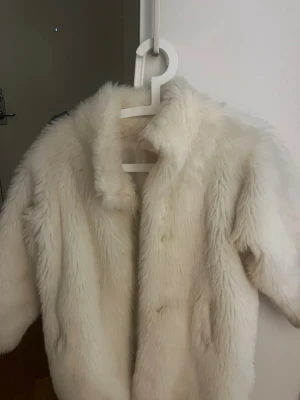 Vit fluffig pälsjacka - Snygg vit pälsjacka med lång ärm och klassisk krage. Jackan är gjord i mjuk fuskpäls som ger en lyxig känsla. Perfekt för att höja din outfit till nästa nivå.