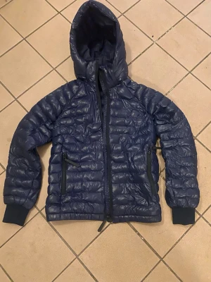 Peak performance Dunjacka - Snygg mörkblå dunjacka med quiltad design och fast huva. Jackan har dragkedja framtill, två sidofickor med dragkedja och muddar vid ärmsluten. Perfekt för kyliga dagar och enkel att matcha med olika outfits.