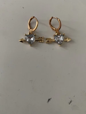 Guldörhängen med hjärtformad sten - Snygga örhängen i guldfärg med pilformad design och en klar hjärtformad sten i mitten. Örhängena har små ringar som fäste och detaljerade pilar som sticker ut på båda sidor om stenen. Perfekta för dig som gillar unika accessoarer.