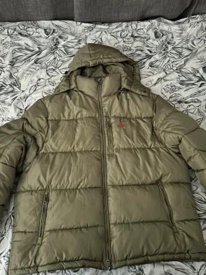 Olivgrön pufferjacka från Polo Ralph Lauren - Säljer en olivgrön pufferjacka från Polo Ralph Lauren med huva och dragkedja framtill. Jackan har två sidofickor med dragkedja och en bröstficka. Klassisk röd Polo-logga på bröstet. Perfekt för kyliga dagar och riktigt snygg streetstil. Storlek XXL ny utan prislapp