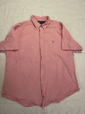 Rosa rutig skjorta från Ralph Lauren - Klassisk rosa och vit rutig skjorta från Ralph Lauren med korta ärmar och button-down krage. Skjortan har broderad logga på bröstet och knappar framtill. Perfekt för en avslappnad men ändå stilren look.
