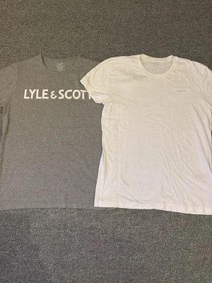 2-pack t-shirts från Lyle & Scott och GANT - Två stilrena t-shirts från Lyle & Scott och GANT, en i grått med stor vit logga över bröstet och en i helvitt med diskret tryck. Båda har rund hals och klassisk passform, perfekta till jeans eller shorts.