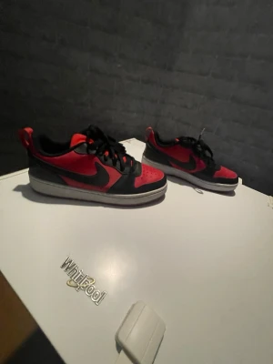 Nike Air Force 1 svart/röd sneakers - Snygga Nike Air Force 1 sneakers i svart och rött med klassisk låg siluett. Skorna har svart swoosh, röd tå och svarta snören. Perfekta för dig som gillar streetstyle och vill sticka ut med färg. Ikonisk design och bekväm passform.