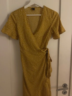 Gul prickig omlottklänning Vero Moda - Superfin gul omlottklänning från Vero Moda med vita prickar. Klänningen har korta ärmar, knyt i midjan och v-ringning.
