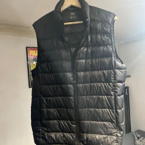 Svart dunväst från Uniqlo Ultra Light Down - Svart, quiltad dunväst från Uniqlo Ultra Light Down. Västen har en stilren, rak passform med hög krage och dragkedja framtill. Lätt och smidig design som är perfekt för lager-på-lager. Materialet är syntet och dun, vilket gör den både varm och lätt.