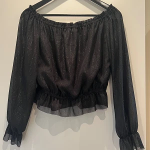 Svart/glittrig off-shoulder blus  - Svart glittrig blus från Divided i storlek 34. Toppen har offshoulder-modell med långa, transparenta ärmar och resår vid axlar, midja och ärmslut. Volangdetalj nedtill ger en extra snygg touch. 