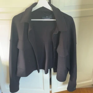 Svart kappa från Stradivarius - Svart kort kappa från Stradivarius med stora bröstfickor och knäppning framtill. Jackan har en loose passform och långa ärmar, perfekt för lager-på-lager. Tjockare material som ger en snygg siluett och trendig look.