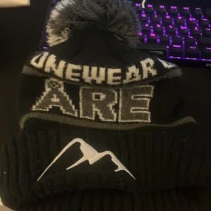Svart Åre mössa med tofs och brodyr - Säljer en svart och grå stickad Åre-mössa från Onewear med stor tofs på toppen. Mössan har broderad vit bergslogga framtill och texten 'ONEWEAR ÅRE' runt om. Perfekt för kalla dagar och vintersäsongen.