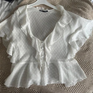 Vit volangblus från Zara - Supersöt vit blus från Zara med volangkant längs v-ringningen och knappar framtill. Tunt, lätt material med diskret prickigt strukturmönster. Kort ärm och peplumliknande nederdel ger en romantisk vibe. Perfekt till jeans eller kjol!