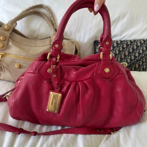 Röd Q Classic Boston Bag! - Perfekta skolväskan! Rymlig röd burgundy Q Classic Boston bag från Marc by Marc Jacobs i äkta läder med guldfärgade detaljer och dubbla handtag. Långa bandet medföljer. Klassisk och tidlös modell som rymmer allt du behöver och passar perfekt både till vardag och resa. Praktiska fack och dragkedja gör den enkel att organisera. Otroligt nyskick, se alla bilder!  Mått 40x22x15 (undersida). Kan hämtas i Nacka eller skickas. Skriv privat för fler bilder 🤍 