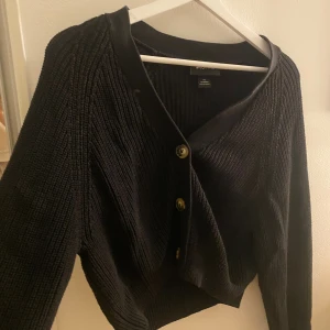 Svart stickad kofta  - Svart stickad kofta från Monki i storlek XS. Koftan har grov ribbad struktur, v-ringad hals och tre snygga knappar framtill. Perfekt att slänga över en t-shirt eller linne för en chill look. Långärmad och lite croppad passform. 