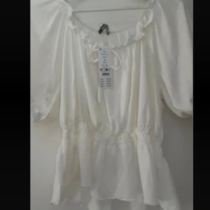 Vit blus med volang och knyt - Supersöt vit blus med volangkant och knyt framtill. Blusen har puffiga korta ärmar och resår i midjan. Kan bäras ut off shoulder.