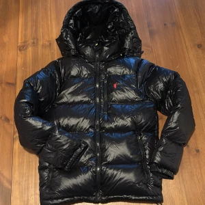 Svart dunjacka från Polo Ralph Lauren - Säljer en svart dunjacka från Polo Ralph Lauren med glansig finish och röd broderad logga på bröstet. Jackan har huva, flera dragkedjeförsedda fickor och quiltad design. Perfekt för kalla dagar och har en riktigt snygg streetstil.