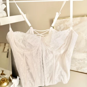 Vit spetskorsett med snörning - Supersnygg vit korsett med spetsdetaljer och justerbara axelband. Baksidan har dekorativ snörning och framsidan stängs med hyskor. Perfekt för att skapa en trendig look med romantisk touch. Fel storlek för mig kan kan inte ändra snörerna 