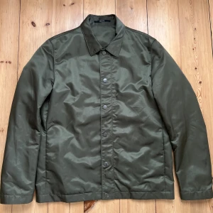 Grön jacka från Tiger of Sweden - Snygg olivgrön vindjacka från Tiger of Sweden med klassisk krage och raka linjer. Jackan har både dragkedja och tryckknappar framtill, samt slät yta i polyester. Perfekt för dig som gillar stilrena och minimalistiska plagg. Vattenavvisande. 