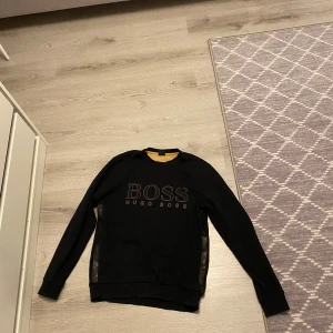 Svart sweatshirt från Hugo Boss - Svart sweatshirt från Hugo Boss med guldfärgade detaljer och stort BOSS-tryck på bröstet. Tröjan har långa ärmar, rund halsringning och dekorativa band med logga längs sidorna. Perfekt för dig som gillar stilren och exklusiv streetwear.