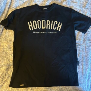 Svart HOODRICH t-shirt med tryck - Svart t-shirt från HOODRICH med stort vitt tryck på bröstet där det står 'HOODRICH' och texten 'FROM NOTHING TO SOMETHING' under. Klassisk passform med rund hals och korta ärmar. Perfekt för en streetwear-look.