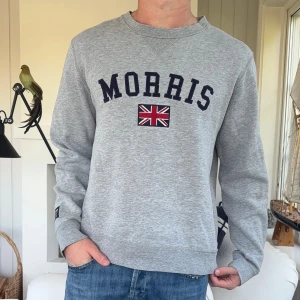 Grå sweatshirt från Morris med tryck - Grå sweatshirt från Morris med mörkblå text och brittisk flagga broderad på bröstet. Klassisk rund halsringning och långa ärmar. Tillverkad i mjuk bomullsmix, perfekt för en avslappnad stil.