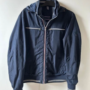 Marinblå vindjacka Tommy Hilfiger - Snygg marinblå vindjacka från Tommy Hilfiger med huva, silverfärgade dragkedjor och broderad logga på ärmen. Jackan har ribbade muddar med vita ränder och två fickor framtill. Perfekt för dig som gillar sportig och stilren look.