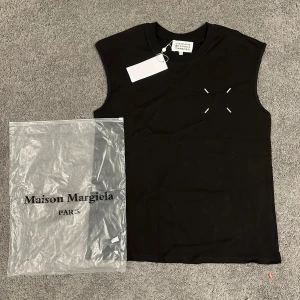 Maison Margiela svart ärmlös topp - Svart ärmlös topp från Maison Margiela med rund hals och vita signatursömmar framtill. Baksidan har ett stort tryck med siffror och logga. Topp i mjuk bomull, perfekt för en clean och edgy look.