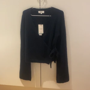 Mörkblå omlottkofta från Gina Tricot - Snygg mörkblå stickad omlottkofta från Selected Femme i storlek S. Koftan har långa ärmar, v-ringad hals och knyt i sidan för en schysst passform. Perfekt att slänga på sig när det är lite kyligt ute.