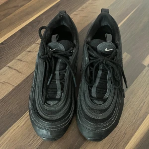 Svarta Nike Air Max sneakers - Säljer ett par svarta Nike Air Max sneakers med klassisk vågig design och synlig luftbubbla i sulan. Skorna har snörning, rund tå och är tillverkade i en mix av syntet och textil. Perfekta för dig som gillar sportig och stilren look.