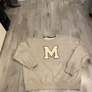 Beige sweatshirt med M från Morris - Snygg beige sweatshirt från Morris med stort broderat M på bröstet. Tröjan har rund hals, ribbade muddar och är långärmad. Perfekt för dig som gillar enkel och stilren design med en sportig touch.