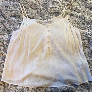 Vit linnetopp med knappar - Säljer en luftig vit linnetopp med smala axelband och knappar framtill. Toppen har en lätt skrynklig struktur. Perfekt för varma dagar och enkel att matcha med olika outfits.Aldrig använd, men har dock ett väldigt litet hål i nederdelen!Kommer med en extra knapp. 