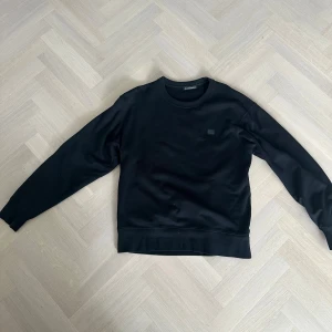 Svart sweatshirt Acne Studios Face L - Svart sweatshirt från Acne Studios i modellen Fairview Face. Tröjan har det ikoniska Face-emblemet broderat på bröstet, rund hals och långa ärmar. Tillverkad i 100% bomull för skön och mjuk känsla. Perfekt för dig som gillar minimalistisk stil.