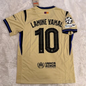 Barcelona Lamine Yamal #10 fotbollströja - Snygg Barcelona fotbollströja från Nike i beige med svarta detaljer och blå inslag. Tryck med Lamine Yamal och nummer 10 på ryggen, samt officiella loggor som Spotify och UNHCR. Kortärmad modell i lätt och ventilerande material, perfekt för match eller träning.