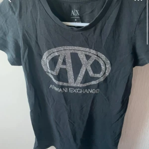 Svart t-shirt Armani Exchange M - Svart t-shirt från Armani Exchange med glittrande AX-logga framtill i silvriga nitar. Klassisk rund halsringning och korta ärmar. Perfekt för en snygg och avslappnad look.😊
