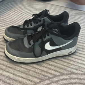Nike Air Force 1 grå och svarta sneakers - Säljer ett par klassiska Nike Air Force 1 sneakers i grått och svart med vit swoosh. Skorna har snörning, låg profil och är tillverkade i skinn och syntet. Perfekta för dig som gillar streetstyle och vill ha en ikonisk look.